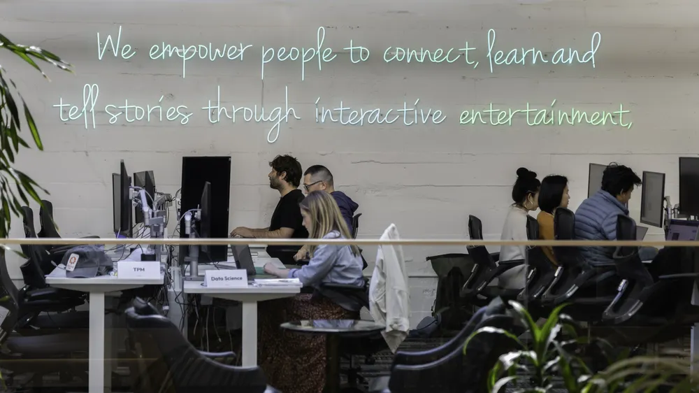 People working at computer stations in an open office space, with a neon sign on the wall that reads 'We empower people to connect, learn and tell stories through interactive entertainment.'