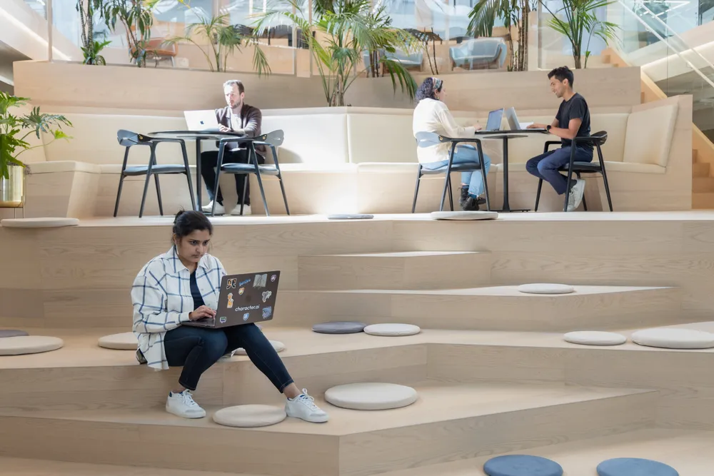 People working on laptops in a modern, open office space with tiered wooden seating and natural light.