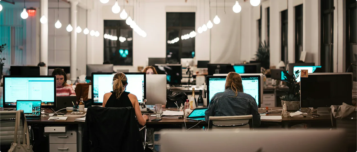 Modern open-plan office at night with several people working at desks equipped with large computer monitors. The room is well-lit with rows of hanging light bulbs and has a minimalistic design featuring white walls, high ceilings, and large windows.
